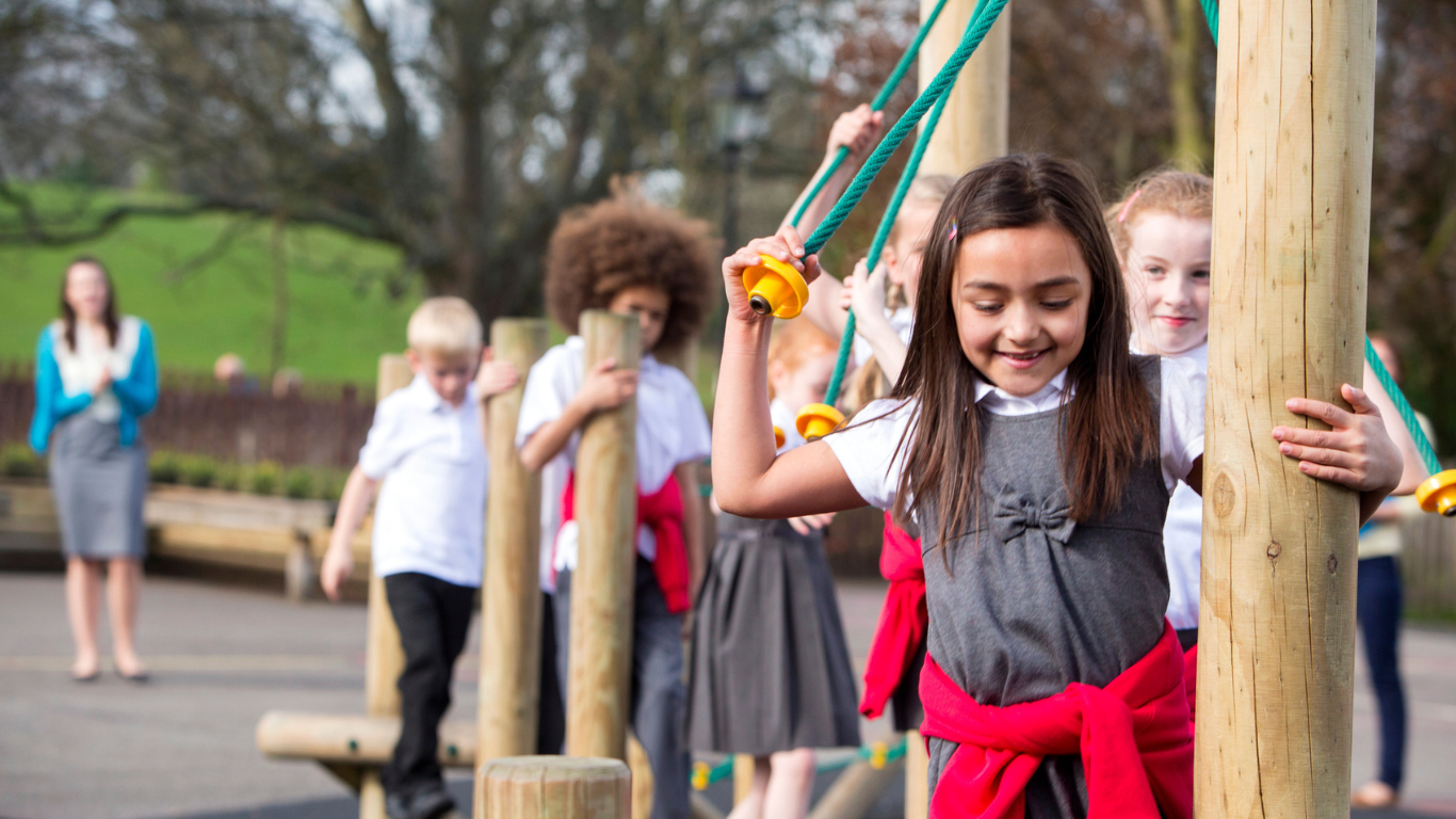 A close up of a group of primary school children playing on a playframe in a playground. Their teacher appears blurred in the background and is supervising. accessible childcare