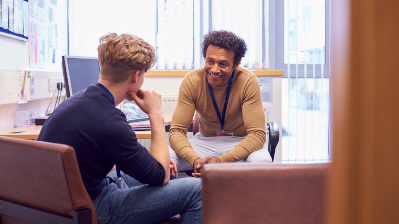 A youth justice practitioner and a young person are sitting at a desk facing each other engaged in a conversation